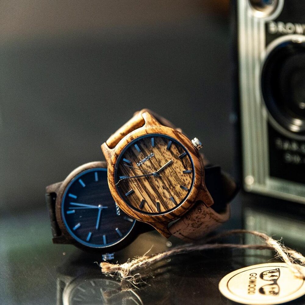 Two wood watches, sit side-by-side on a glass table. The watches are made from natural wood, which is often associated with love and romance