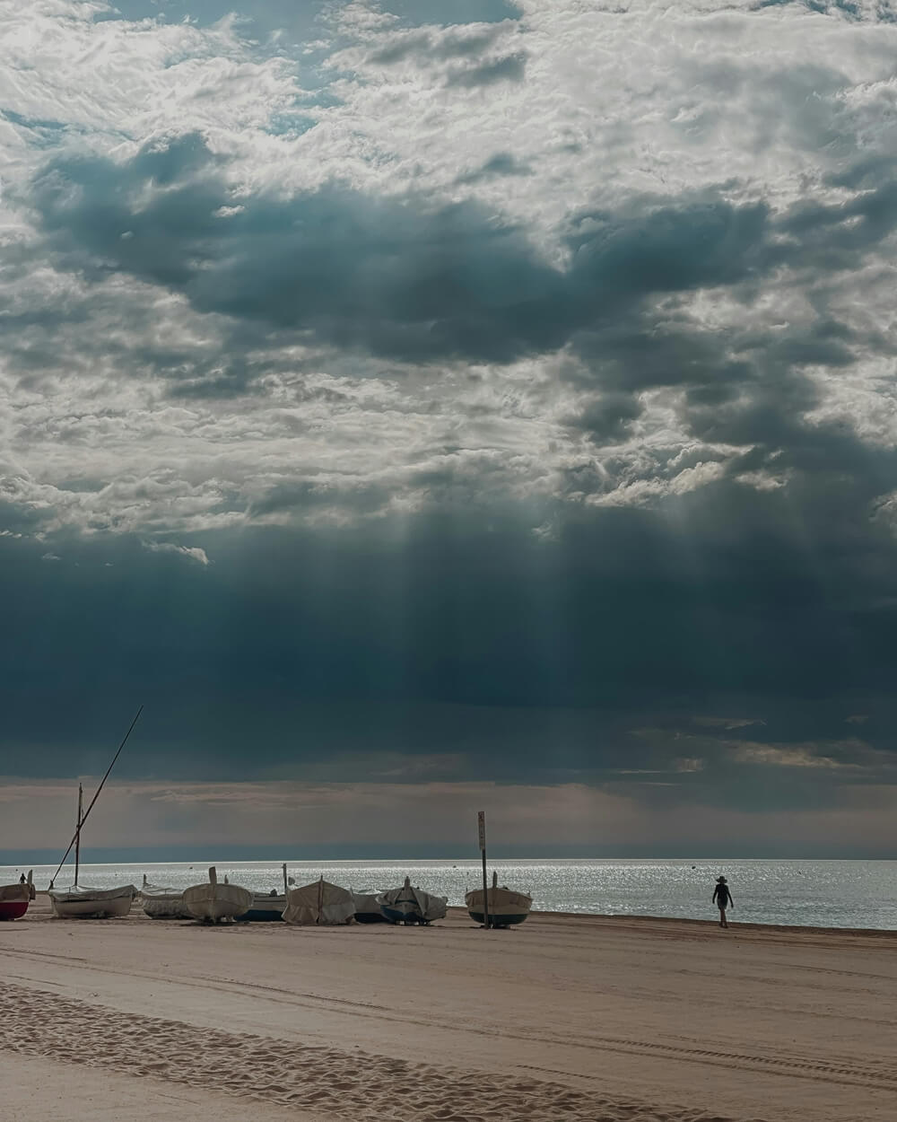 A cloudy sky with sun rays breaking through over a calm ocean and a sandy beach where several small boats are pulled ashore
