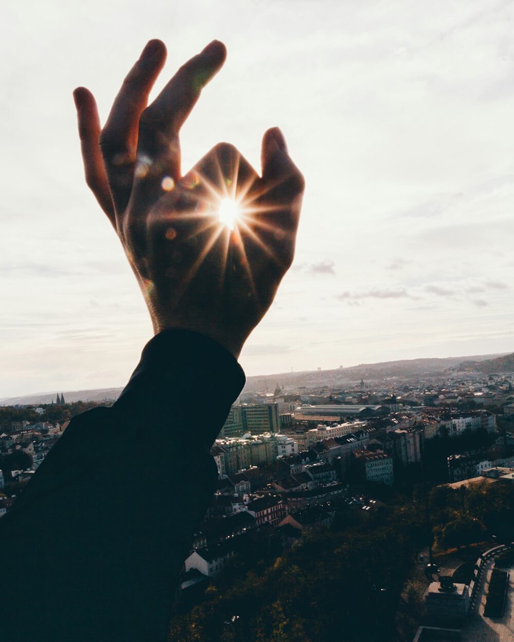 A hand held up to the sky, with the sun shining brightly through the circle made by the thumb and index finger, above a city skyline