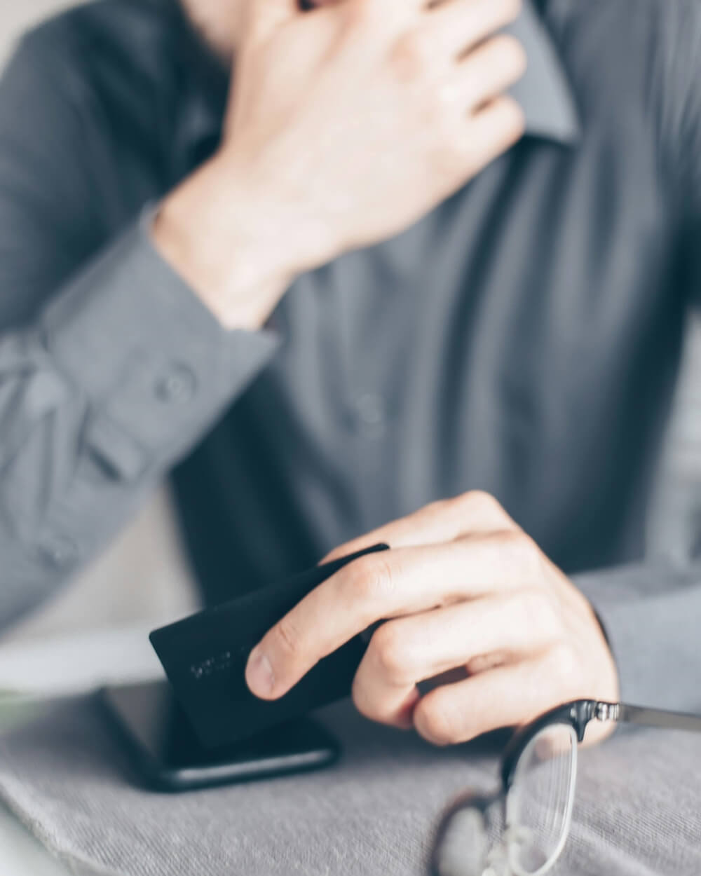 A person holding a black credit card above a smartphone, with eyeglasses resting on a grey surface