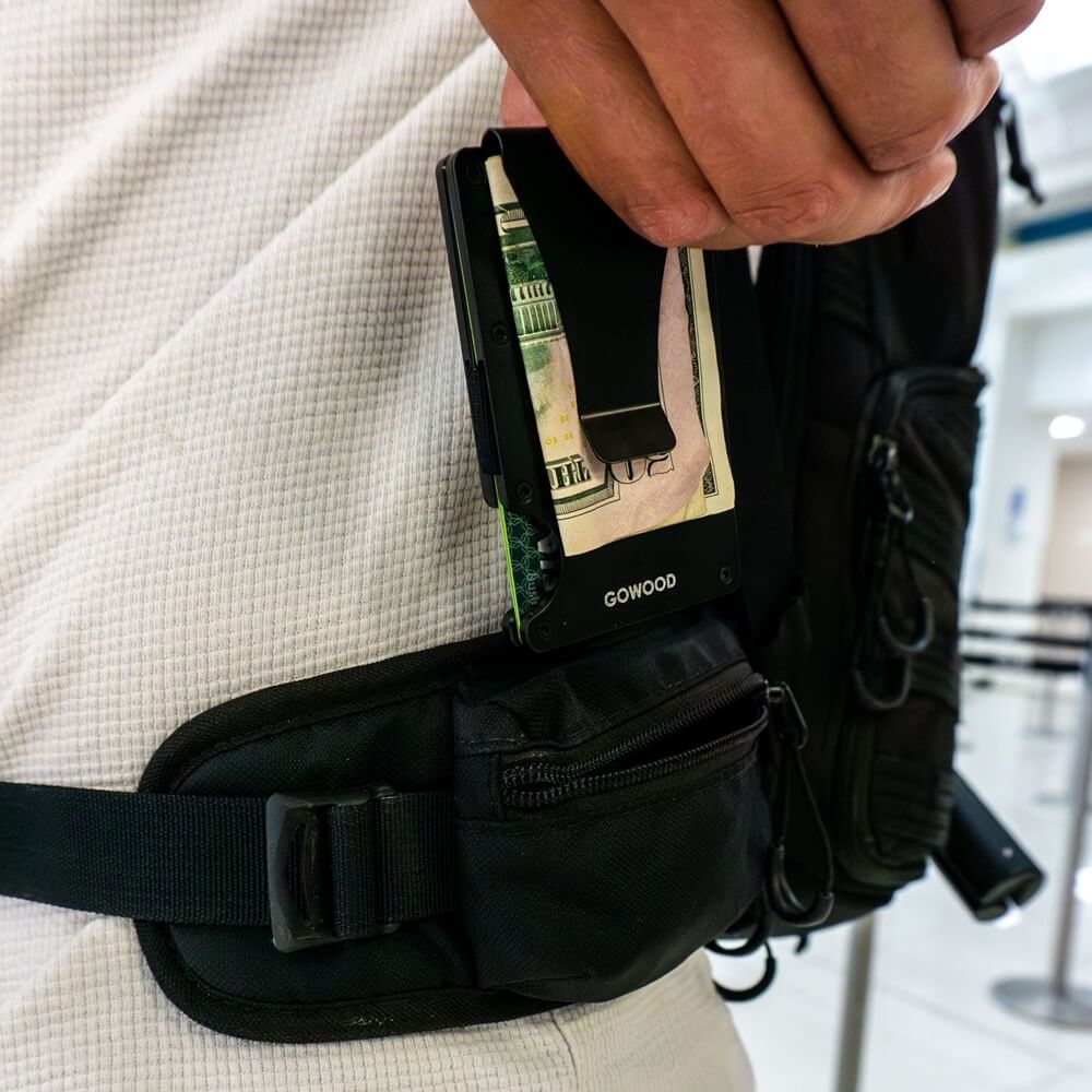 A person holding a black minimalist RFID-blocking wallet with money inside, near a black fanny pack or bag