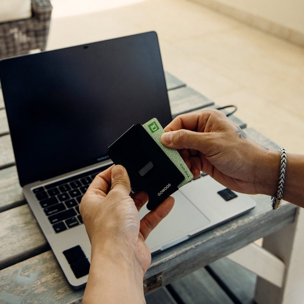 A person's hands holding a black minimalist wallet with a green card visible, positioned above an open laptop on a wooden table