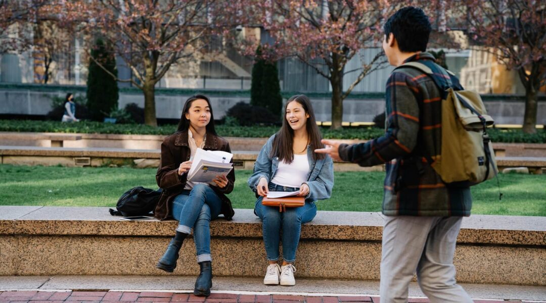 Two students are sitting on a college campus and another one is holding a book, the scene captures a back-to-school atmosphere, where a key accessory could be a wooden watch