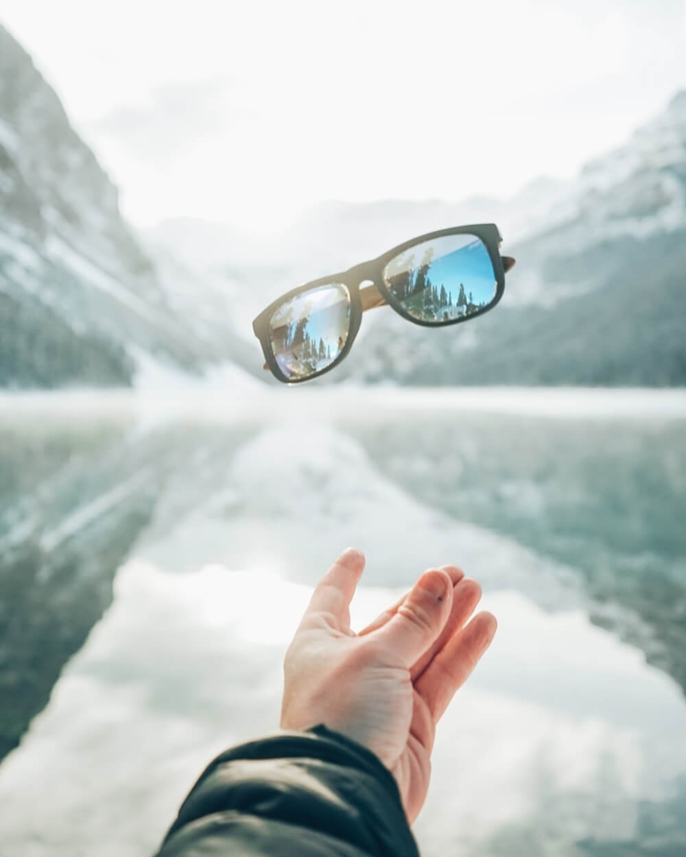 A hand reaches up to a pair of Gowood wooden sunglasses, which reflect a crisp, snowy mountain and pine tree landscape mirrored in a serene lake below