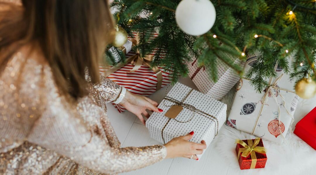 A woman in a sparkly gold dress places an eco-friendly holiday gift wrapped in white paper with a brown tag and twine under a Christmas tree