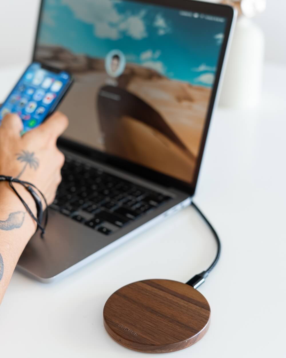 A person with tattoos using a laptop and holding a phone, with wooden wireless chargers